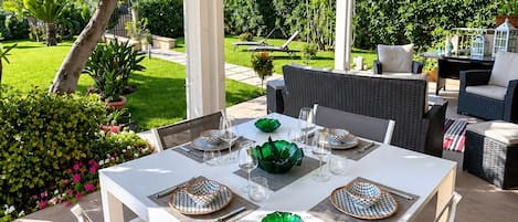 An outdoor patio with a white table set for dining, surrounded by greenery and a view of a well-maintained garden.