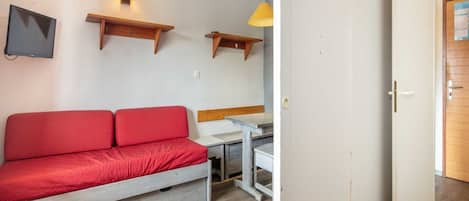 Ein Zimmer mit einem roten Sofa, einem kleinen Tisch, einem an der Wand befestigten Fernseher und einer Tür.