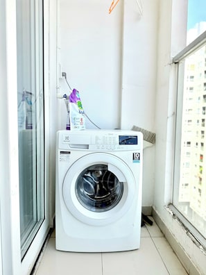 Eine Waschmaschine mit Frontlader in einem kleinen, abgeschlossenen Balkonbereich mit Fenster und Wandrohr.