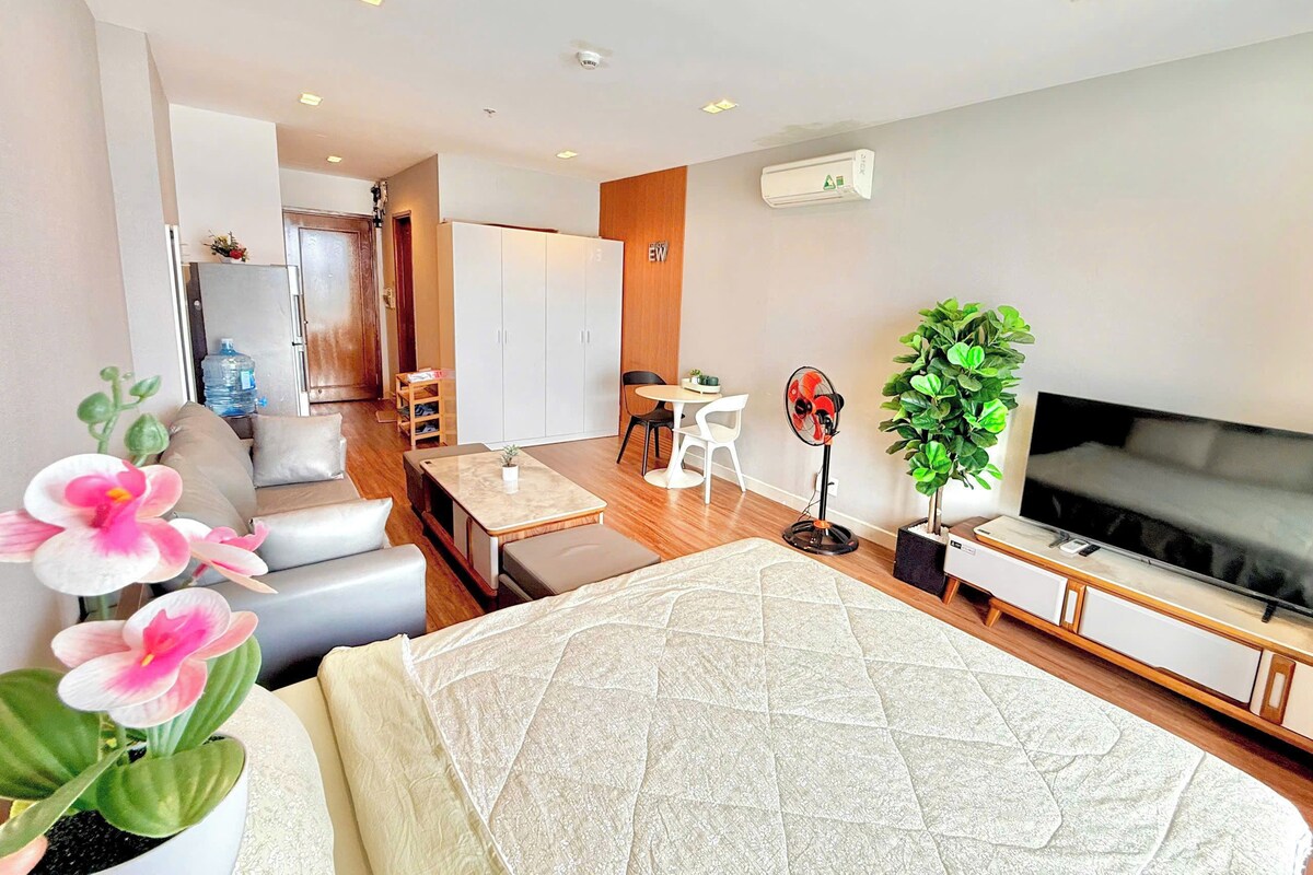 Ein modernes Hotelzimmer mit Bett, Fernseher, Schreibtisch, Sessel, Sofa, Pflanze und Wasserspender.