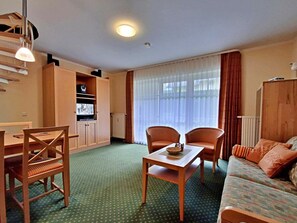 Ein kompaktes Hotelzimmer mit grüner Teppichböden, einer Couch, einem Esstisch mit Stühlen, einem Fernseher auf einem Schrank und einem Fenster mit Vorhängen.