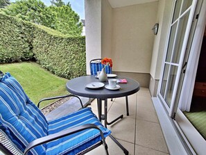 Eine kleine Terrasse mit einem runden Tisch und Stühlen, einem Blumenstrauß in einer Vase und Blick auf einen Hecken- und Rasenbereich.