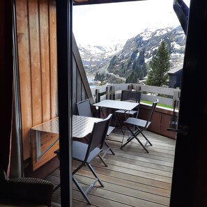 Una terraza de madera con mesas y sillas, con vistas a un paisaje montañoso con picos nevados y un lago.
