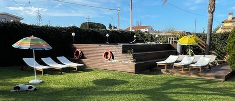 A backyard with a grassy lawn, a wooden deck, and a pool area with lounge chairs and umbrellas.