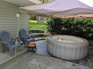 Un patio al aire libre con un hogar para fuego, dos sillas Adirondack y un jacuzzi redondo grande bajo una marquesina.