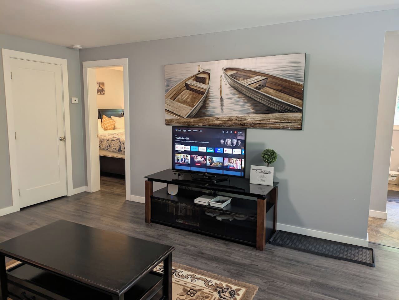 A living room with a flat-screen TV on a black stand, a coffee table, and a wall-mounted painting of boats.