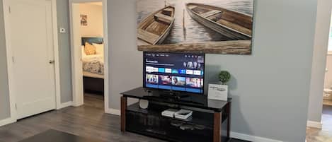 A living room with a flat-screen TV on a black stand, a coffee table, and a wall-mounted painting of boats.