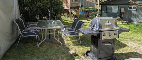 An outdoor patio with a glass table, metal chairs, a barbecue grill, and a playhouse in the background.
