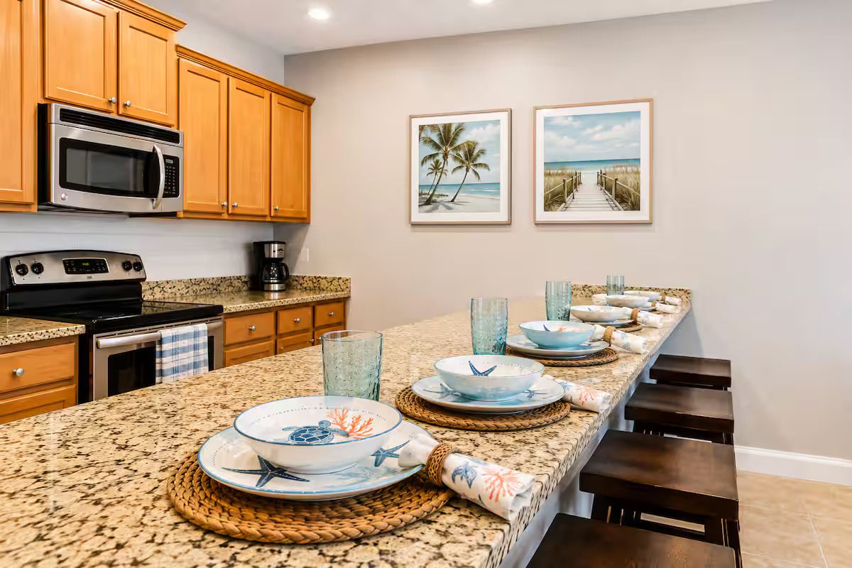 A kitchen with wooden cabinets, a microwave, a stove, and a granite countertop. There are framed pictures on the wall and a long dining table set with plates and glasses.