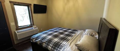 Un dormitorio con una cama, una ventana, un televisor montado en la pared y un ventilador de techo.