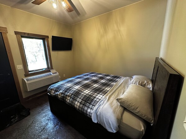 A bedroom with a bed, a window, a wall-mounted TV, and a ceiling fan.