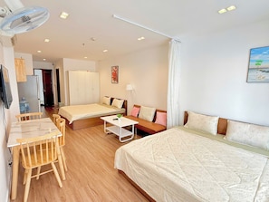 A hotel room with a bed, a sofa, a desk with chairs, a standing fan, and a painting on the wall. - AIGO LUXURY APARTMENT (ho chi minh)