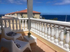 A balcony with white chairs and a railing, overlooking a coastal view.