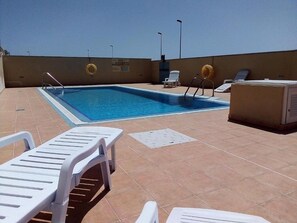 A rooftop pool area with a clear blue pool, white lounge chairs, and a small seating area.
