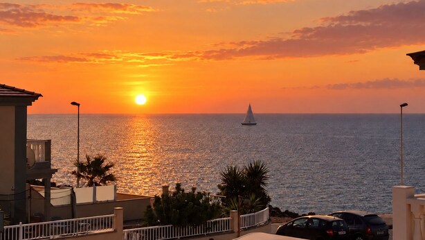 A sunset over the sea with a sailboat, buildings, and a car in the foreground.