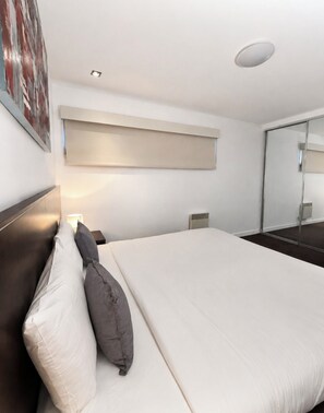 Ein modernes Hotelzimmer mit einem großen Bett, einem Kopfteil, einem Nachttisch, einer Wandleuchte und einer Deckenleuchte sowie einem großen Spiegel.