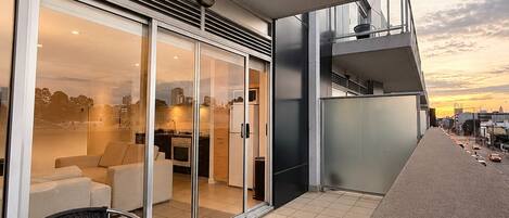 A modern outdoor balcony with a glass door leading to a kitchen, a round table with chairs, and a view of the cityscape.