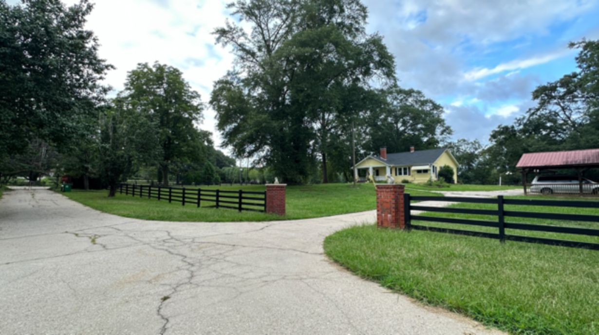 A paved driveway leading to a house with a fenced yard and a covered structure.