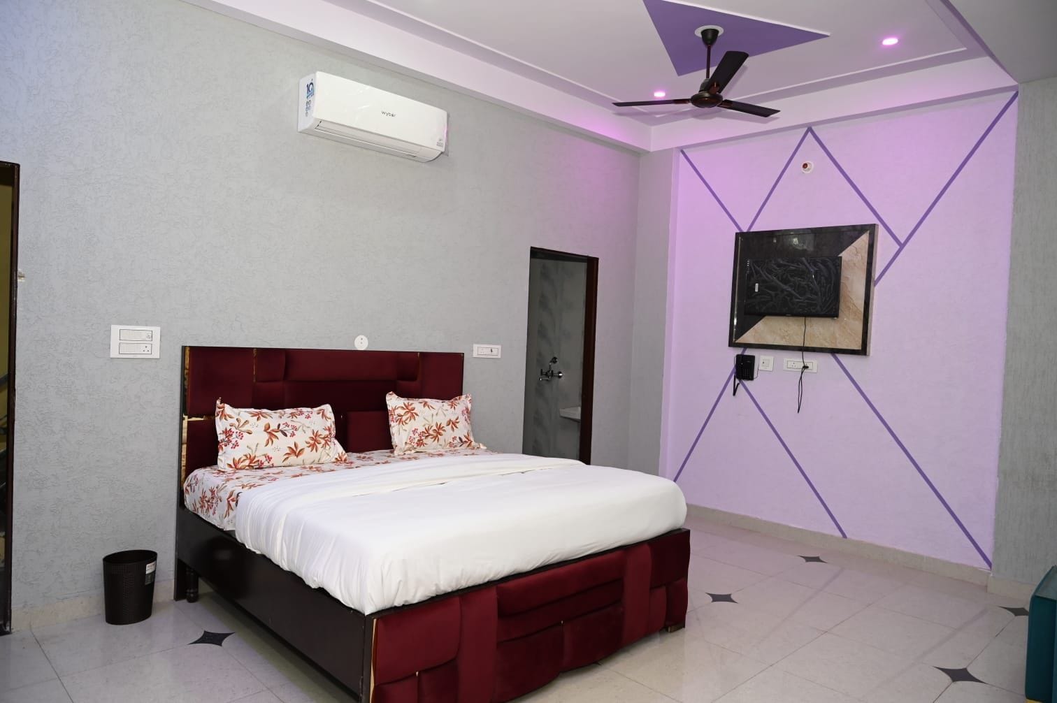 Un dormitorio con cama, ventilador de techo, unidad de aire acondicionado, televisor montado en la pared y una puerta.
