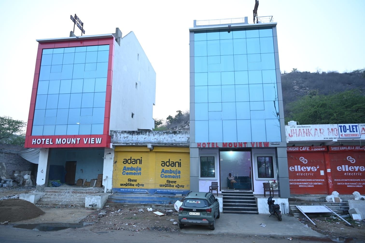 A hotel named 'Hotel Mount View' with a red and white facade, a yellow signboard for 'Adani Ambuja Cement,' and a 'To-Let' signboard.