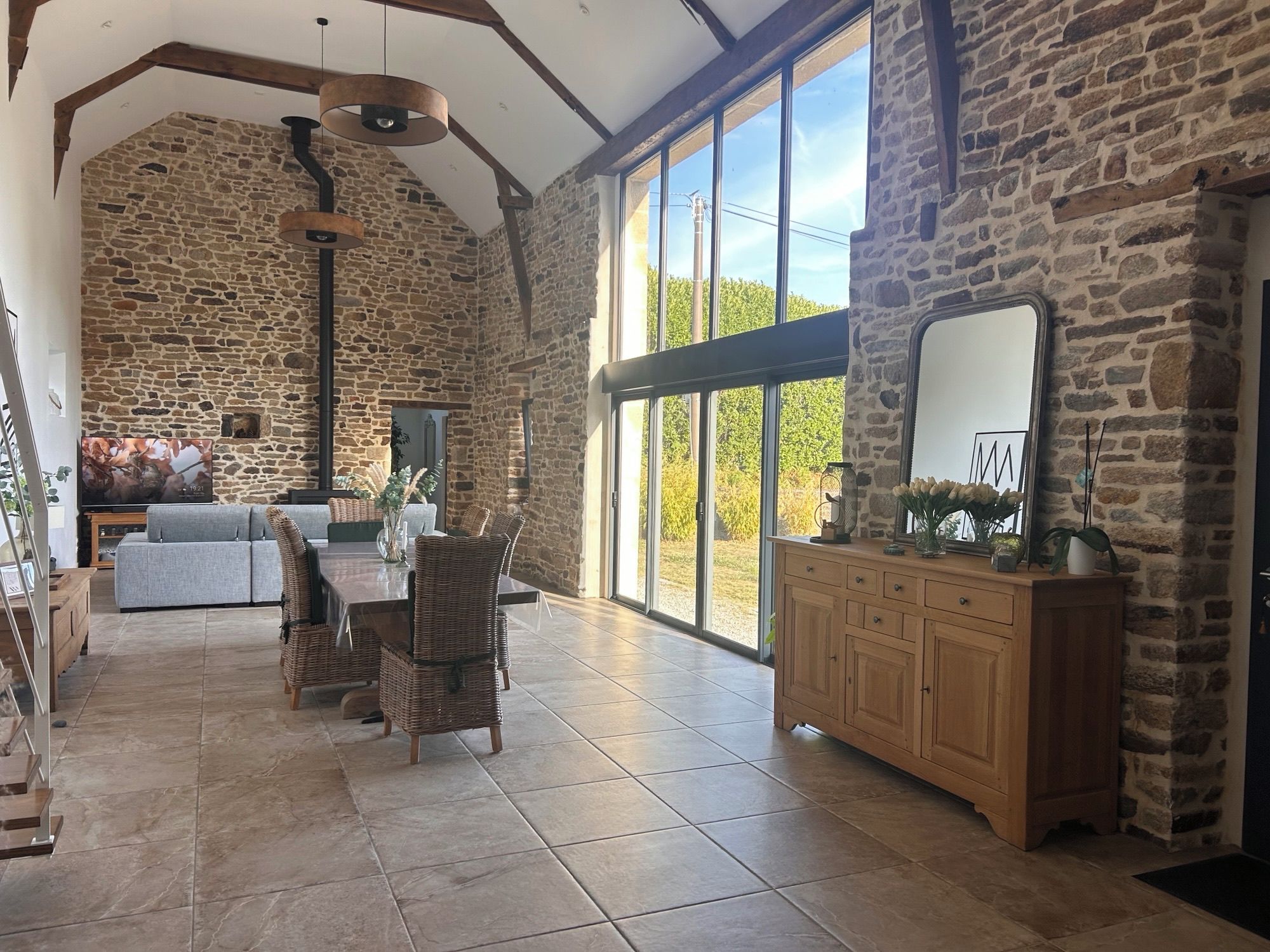 A spacious room with a stone wall, a large window, a dining table with chairs, and a wooden cabinet.