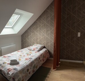 Ein Schlafzimmer mit geneigter Decke, einem Oberlicht, einem Bett mit floralem Bettzeug, einer Wand mit Muster und einem Vorhang.