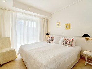 Ein Hotelzimmer mit einem großen Bett, einem Sessel, zwei Wandlampen und einem Fenster mit durchsichtigen Vorhängen.