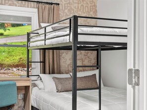 A bunk bed with a desk and chair, a window with curtains, and a view of greenery outside.