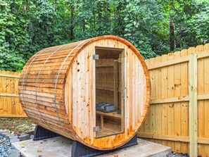A cylindrical wooden sauna structure with a glass door, set on a concrete base, surrounded by a wooden fence and greenery.