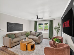 A modern living room with a sectional sofa, a round wooden coffee table, a television, and a large window with curtains.