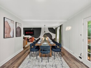 A dining area with a long table set for six, flanked by two sofas and a TV screen displaying 'NETFLIX'.