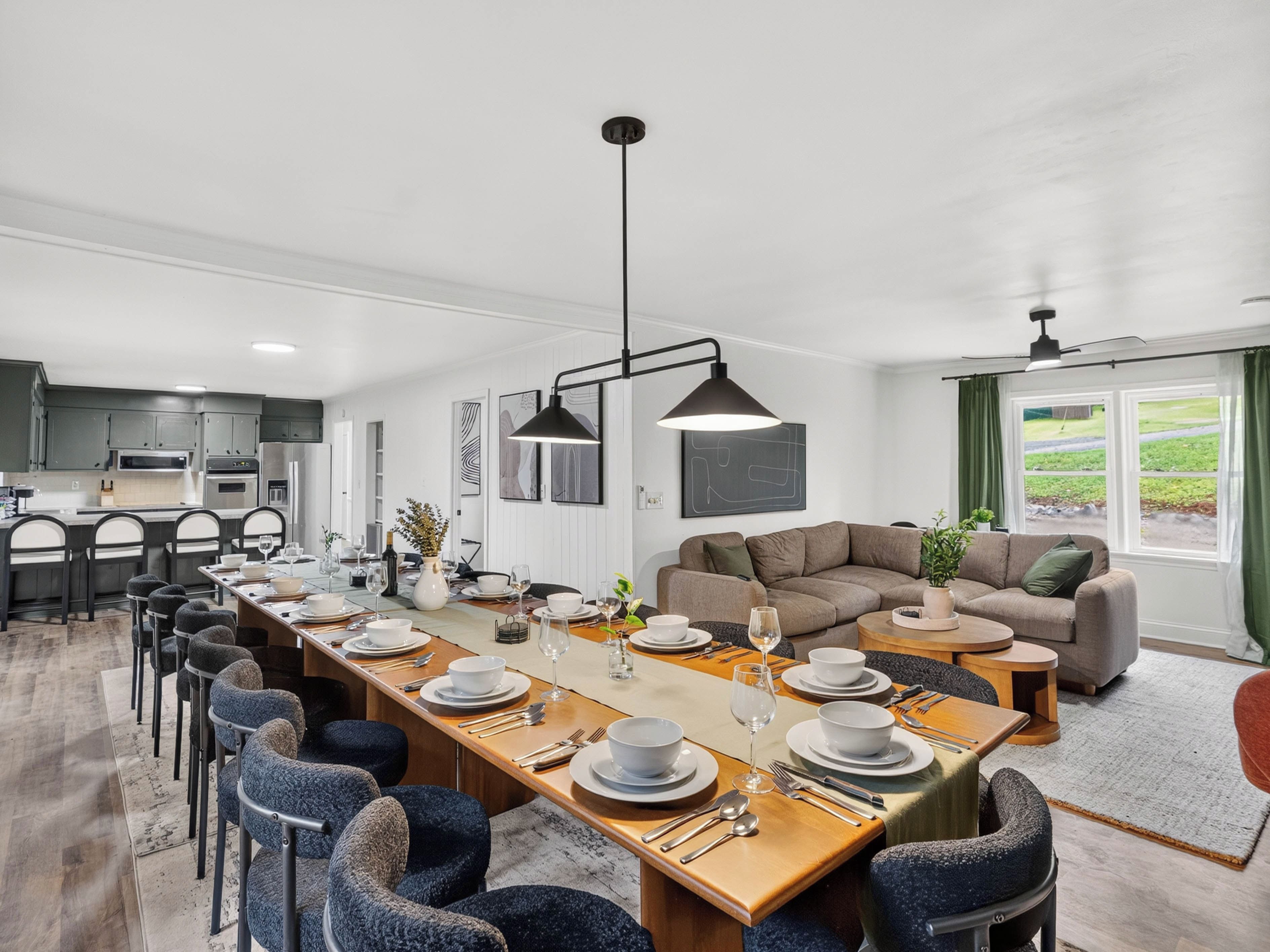 A modern kitchen with dark cabinets, a dining area with a long table set for six, and a living area with a sofa and a window with curtains.