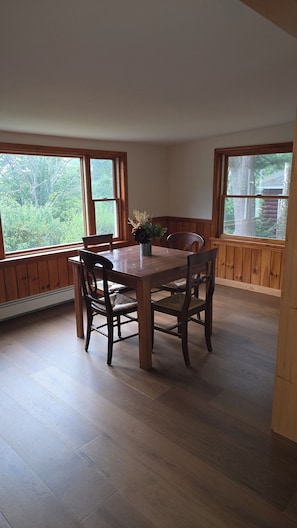 Comedor con mesa y sillas de madera, ventanas amplias y vista a árboles.