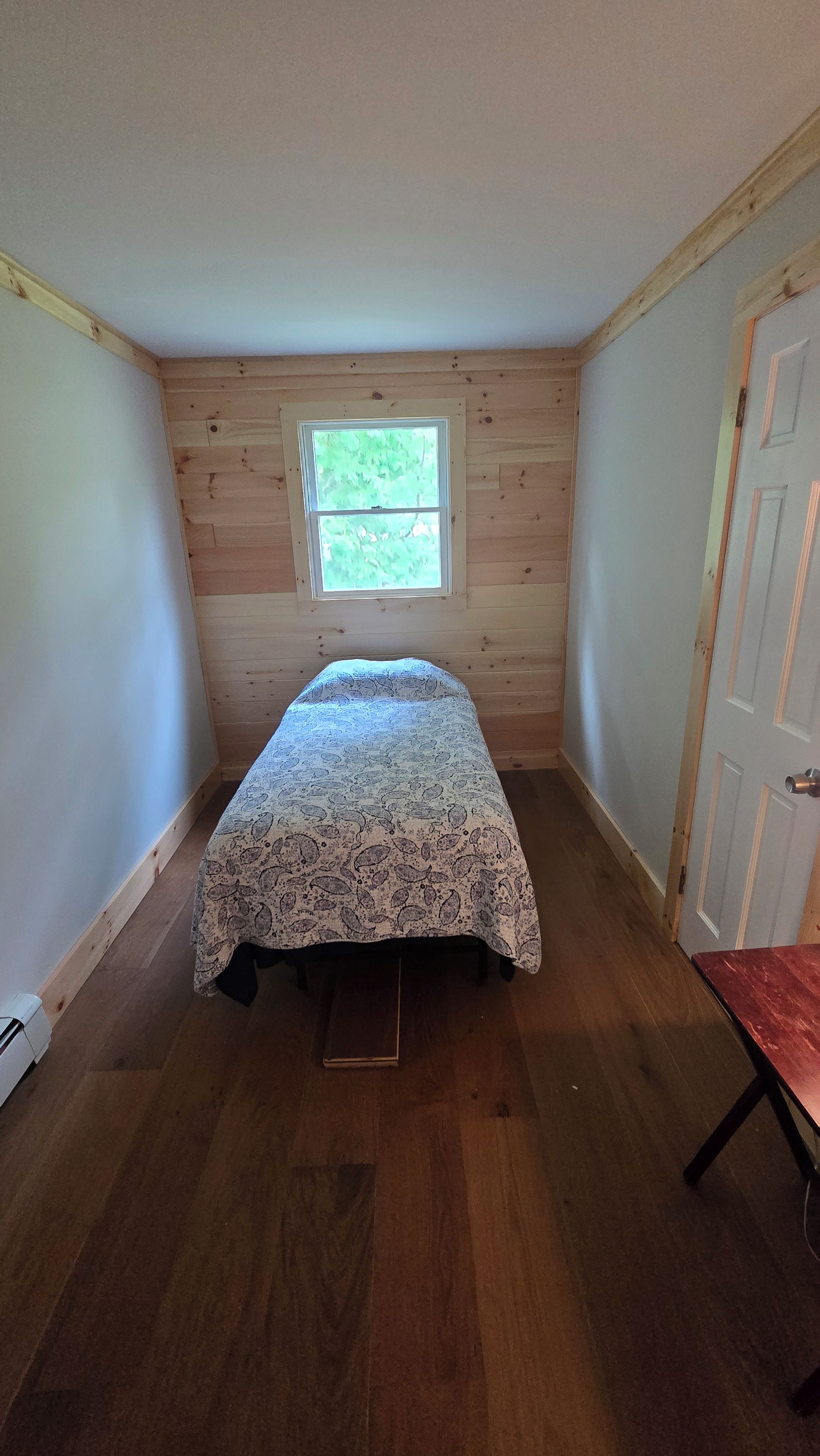 Ein kleines Zimmer mit einem Einzelbett, Holzwänden, einem Fenster und einer Tür.
