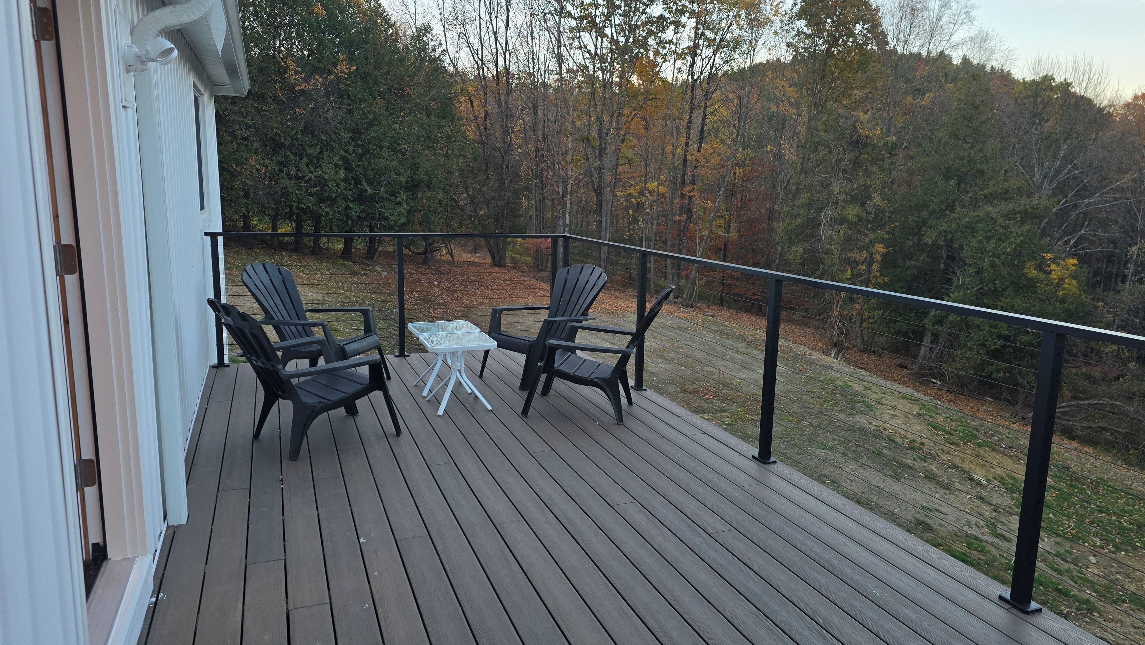 Eine Holzterrasse mit zwei Adirondack-Stühlen und einem kleinen Tisch, mit Blick auf eine bewaldete Landschaft.