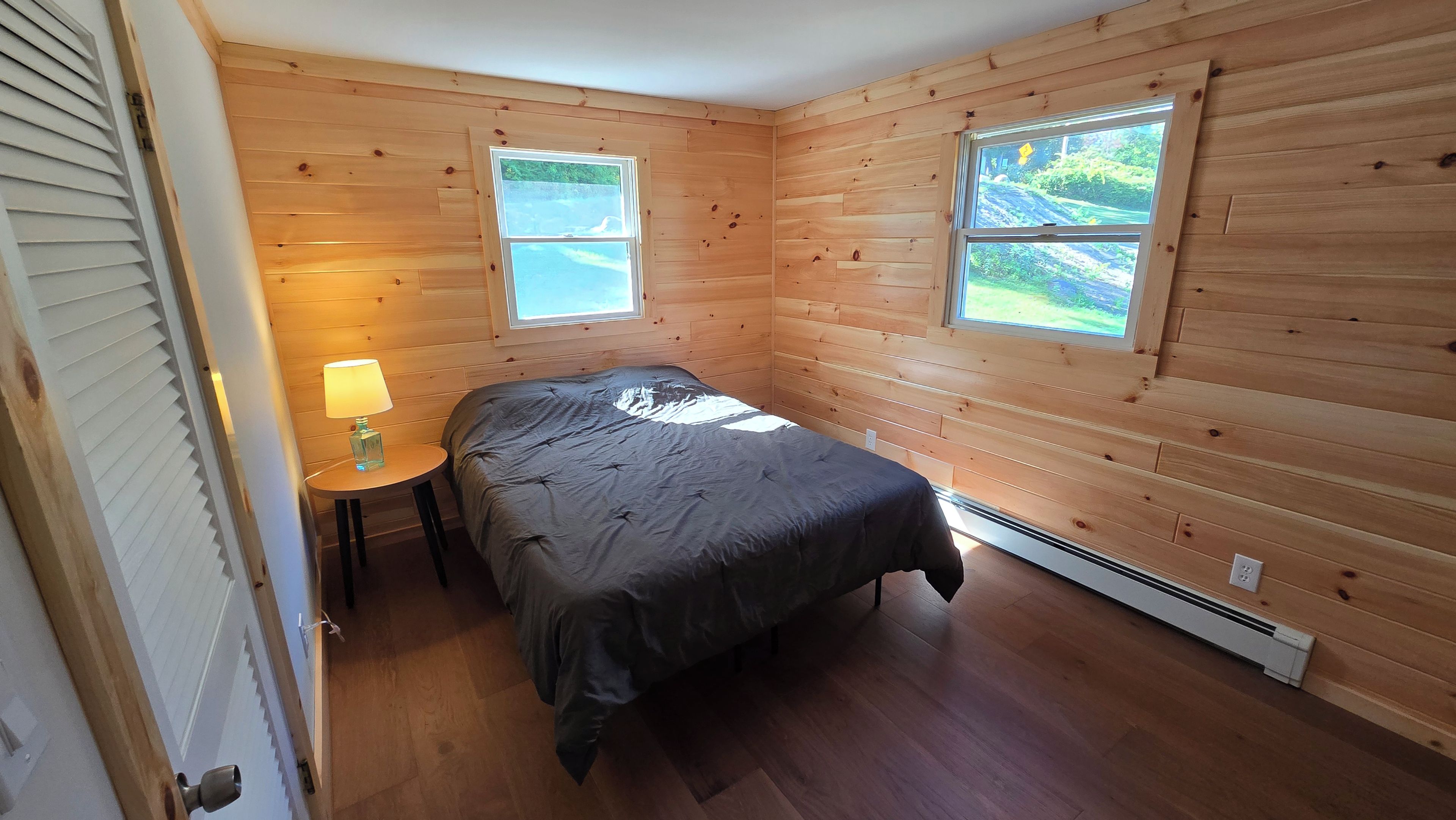 A bedroom with wooden walls, a bed, a nightstand, a lamp, and two windows.