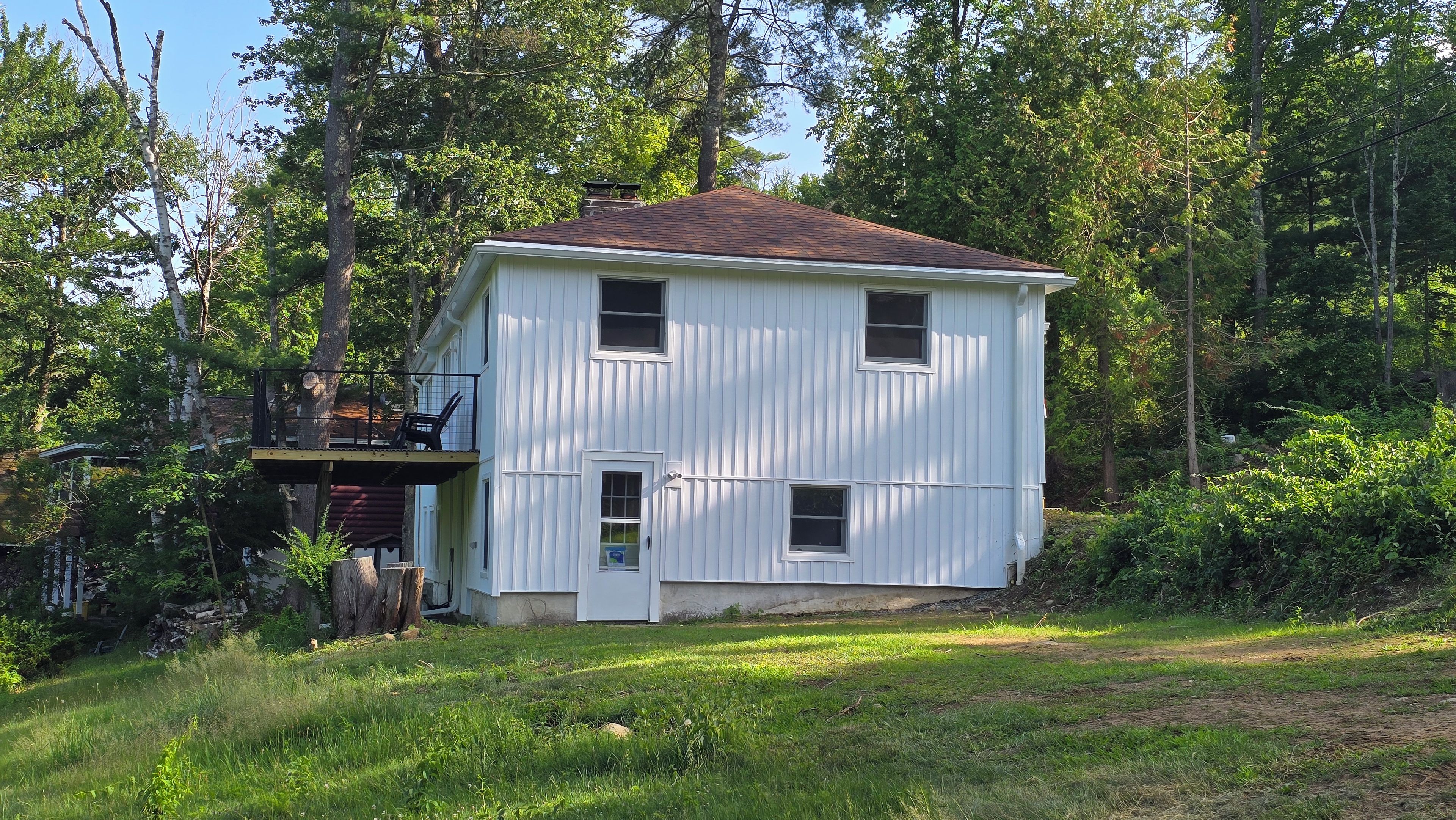 A two-story white wooden house with a balcony, surrounded by trees and a grassy area.