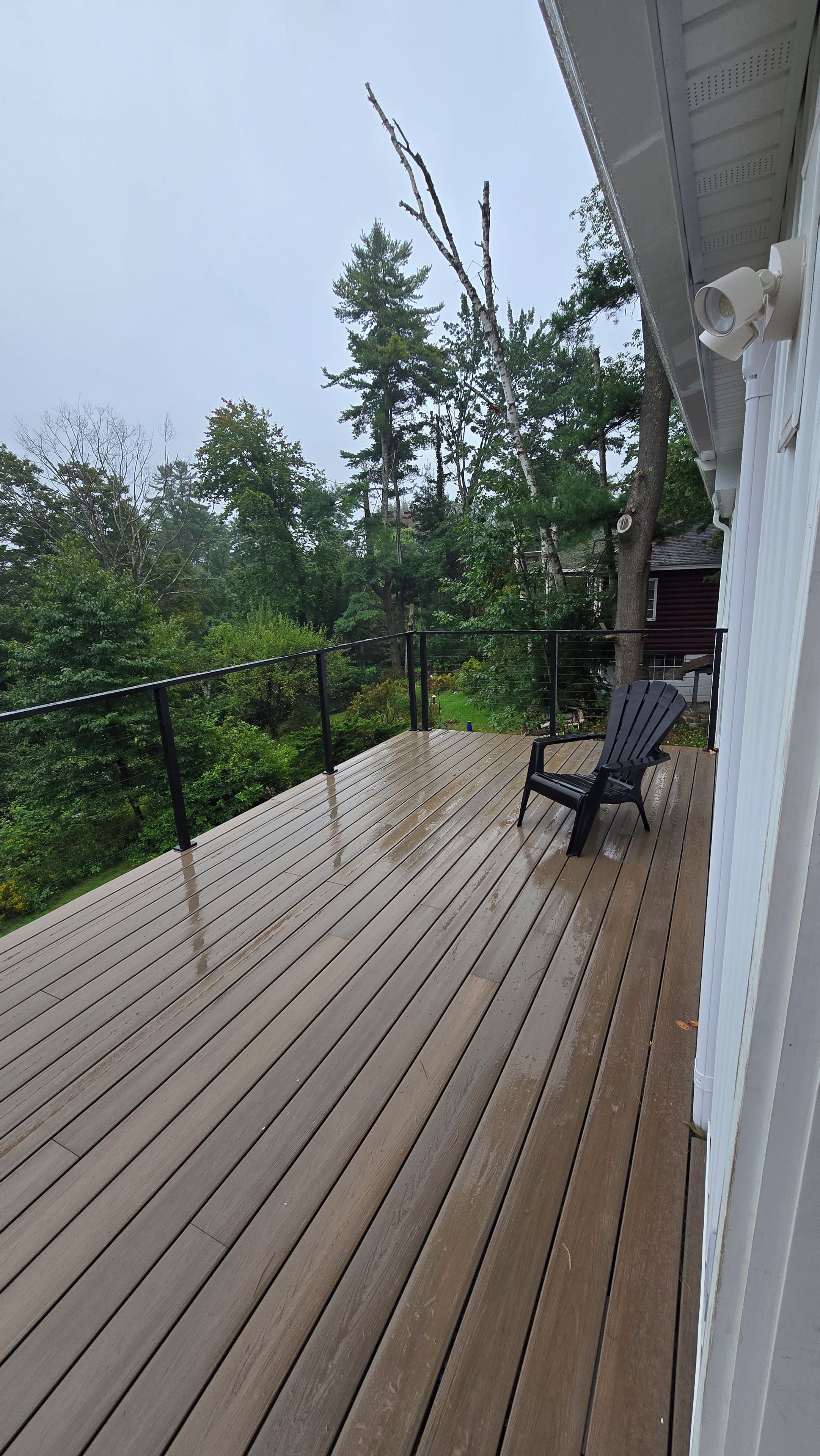 Une terrasse en bois avec une rambarde, une chaise noire et une vue sur des arbres et une maison au loin.