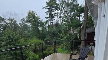 Een houten terras met een leuning, een zwarte stoel en een uitzicht op bomen en een huis op de achtergrond.