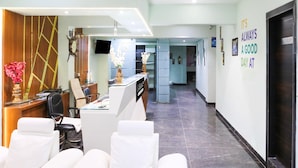 Een hotel lobby met een receptiebalie, een zithoek met witte banken, een klok en een bord aan de muur.