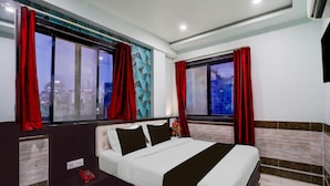 Een moderne hotelkamer met een groot bed, twee ramen met rode gordijnen, uitzicht op de stad en een klein nachtkastje met telefoon.