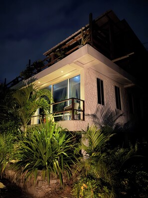 A modern house with large windows and a balcony, surrounded by lush greenery.