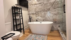 Un baño moderno con una bañera independiente, un inodoro, un lavabo y un televisor montado en la pared, además de una alfombra estampada en el suelo.