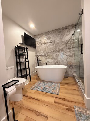 Een moderne badkamer met een vrijstaand bad, toilet, wastafel, een aan de muur bevestigde televisie en een vloerkleed met een patroon.
