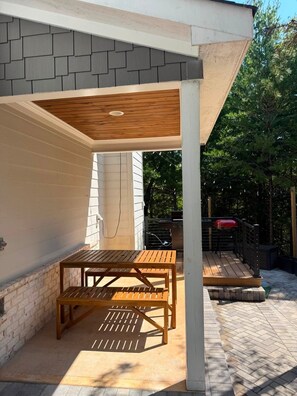 Een buitenpatio met een houten tafel en stoelen, een overdekt gedeelte met een wit plafond en een bakstenen muur.