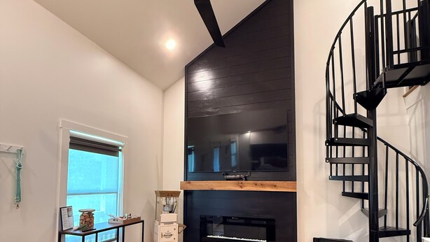 Una sala moderna con una escalera espiral negra, ventilador de techo, chimenea y un área de descanso con otomanas.