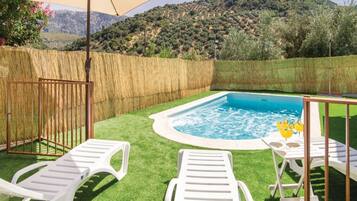 A backyard with a swimming pool, two white lounge chairs, a table with drinks, and a view of a hilly landscape.