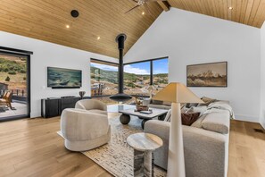 A spacious living room with a high ceiling, a fireplace, comfortable seating, and large windows offering a view of the countryside. - Wasatch View - Luxury Alpine Retreat with Private Hot Tub and Mountain Views (Daniel)