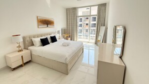 A modern hotel room with a large bed, a bedside table with a lamp, a vanity with a mirror, and a view of the city through the window. - Bright and Modern One Bedroom Apartment (Arjan-Dubailand)
