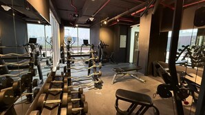 A gym with various exercise equipment, a bench, and a view of the city through large windows. - Bright and Modern One Bedroom Apartment (Arjan-Dubailand)